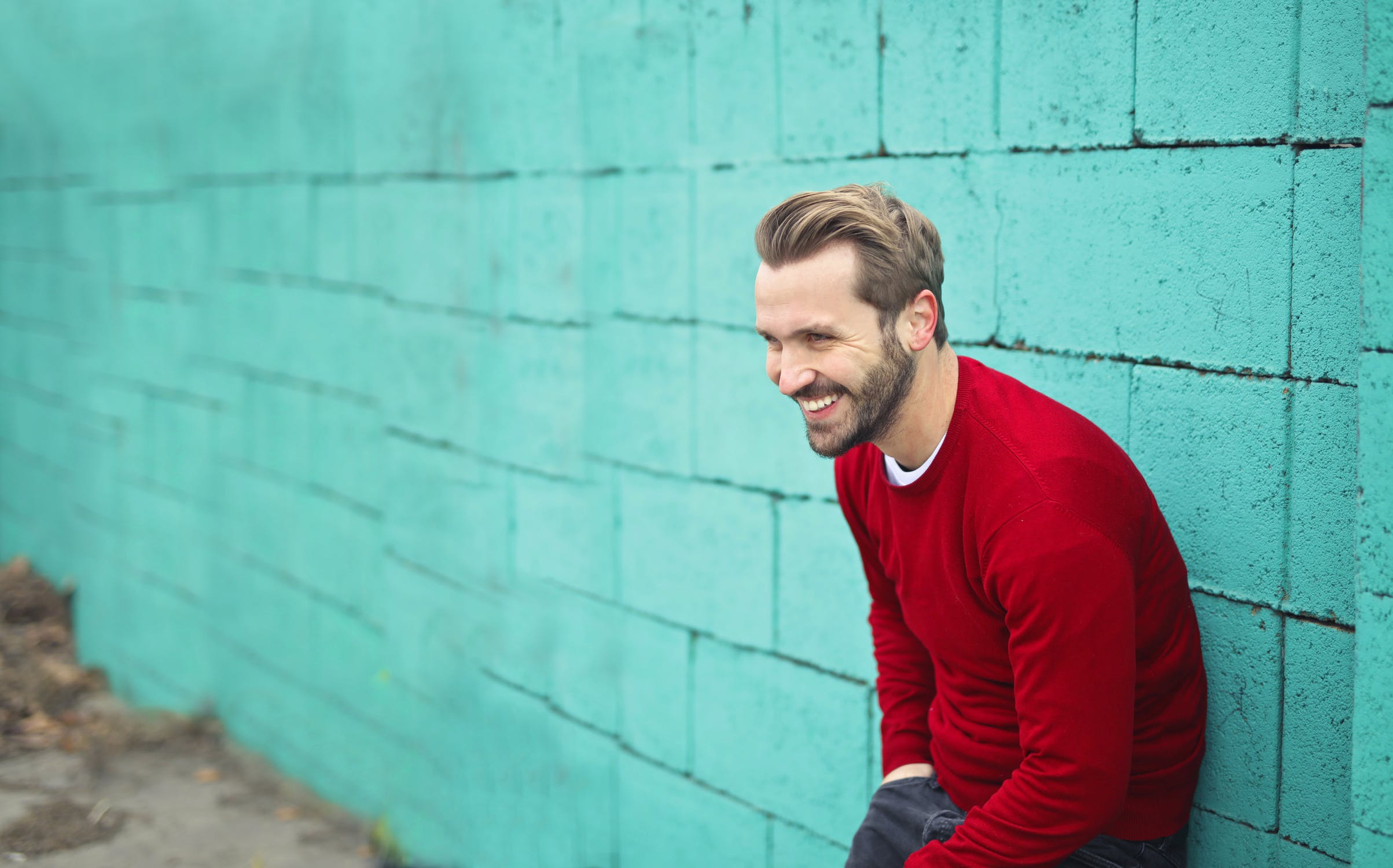 A man in a red jumper leaning against a teal wall, smiling
